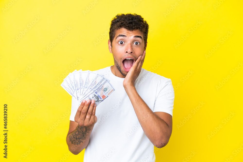 Young brazilian man taking a lot of money isolated on yellow background with surprise and shocked facial expression Young brazilian man taking a lot of money isolated on yellow background with surprise and shocked facial expression