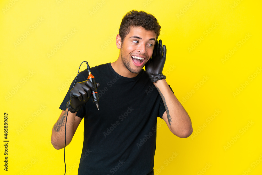Tattoo artist Brazilian man isolated on yellow background listening to something by putting hand on the ear Tattoo artist Brazilian man isolated on yellow background listening to something by putting hand on the ear