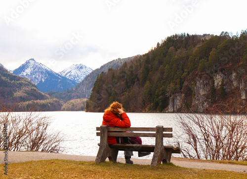 Adult woman sitting on a wooden bench, admiring a snowy mountain landscape | Scene of Alpsee lake in the alps in Bavaria of southern Germany, Europe