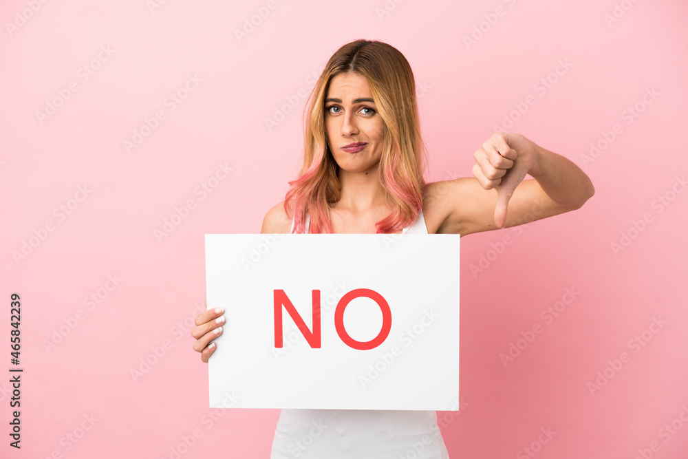 Young woman over isolated pink background holding a placard with text NO and doing bad signal Young woman over isolated pink background holding a placard with text NO and doing bad signal