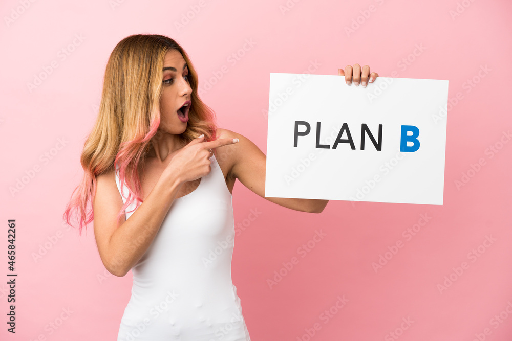 Young woman over isolated pink background holding a placard with the message PLAN B with surprised expression Young woman over isolated pink background holding a placard with the message PLAN B with surprised expression