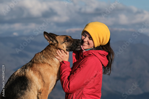 Hug and kiss best friend in nature. Young pretty Caucasian woman in red jacket stands on top of mountain next to her dog and enjoys scenery at sunset. Travel with German shepherd and owner. © Ekaterina