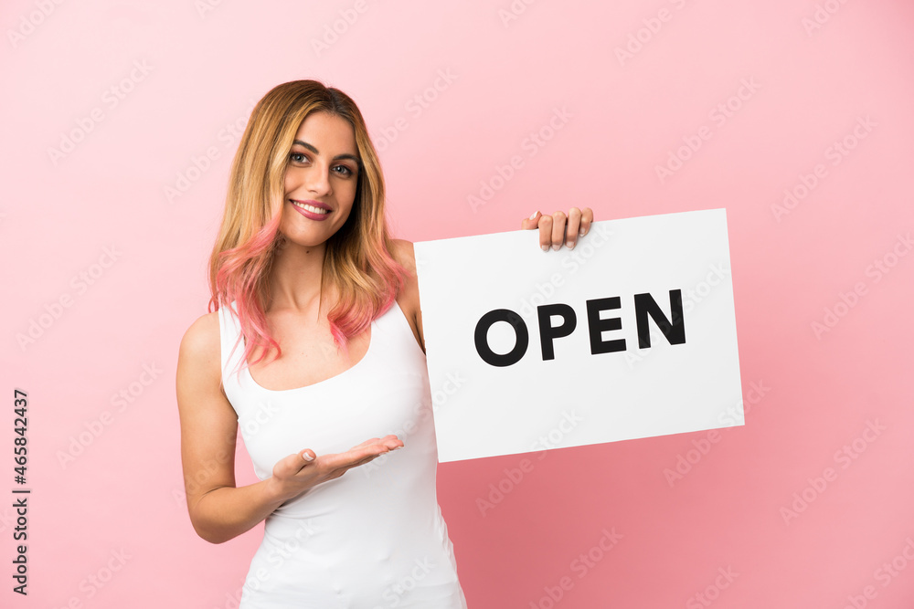 Young woman over isolated pink background holding a placard with text OPEN and pointing it Young woman over isolated pink background holding a placard with text OPEN and pointing it