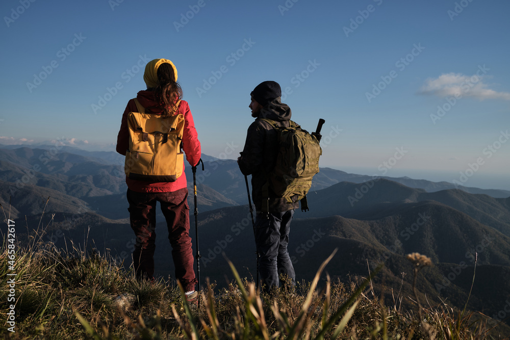 Travel in mountains with loved one and enjoy beautiful panoramic view. Man and woman travelers in red jacket with yellow backpack are standing with their backs turned and looking at mountains.