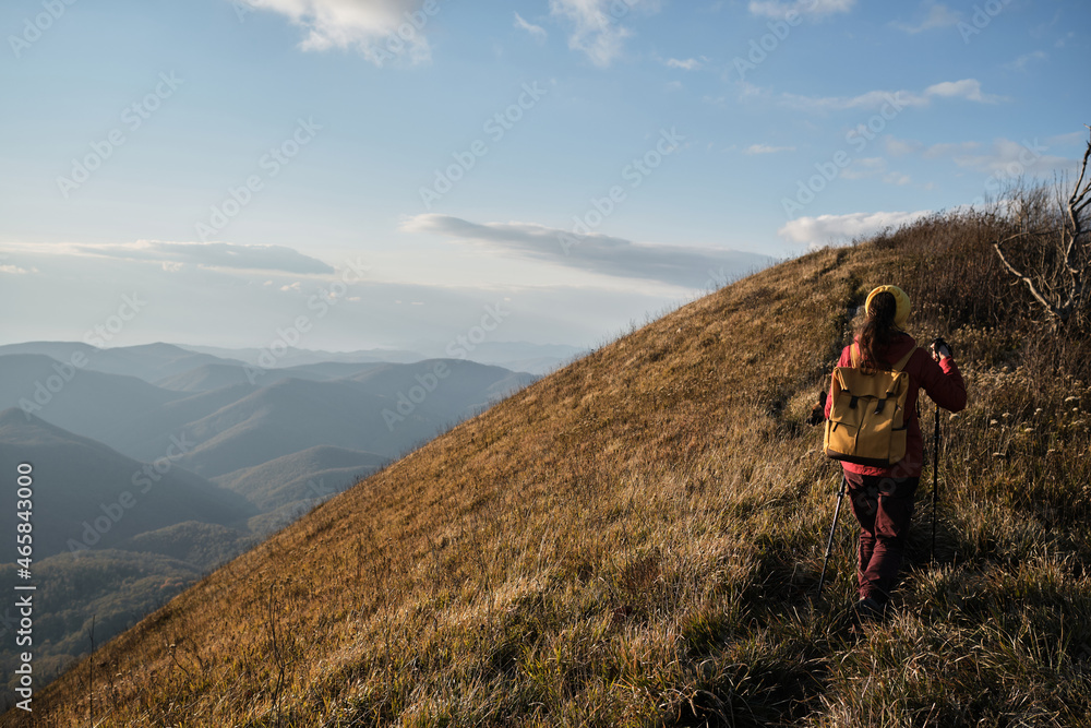 Trekking route in national park. Walk above clouds with backpack and sticks. Female traveler walks along narrow path on top of mountain among tall yellow dry grass.