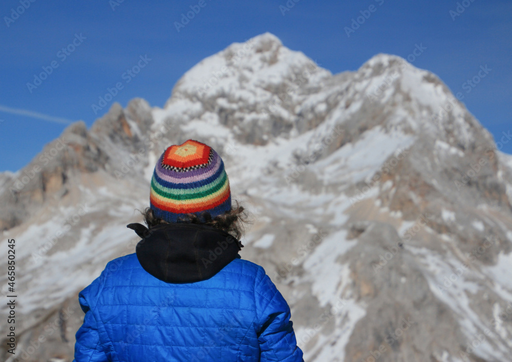 Rear view of  woman with colorful woll hat standing at the top of a mountain, looking into the distance on the top of mountain. Background is mountains and clear sky.