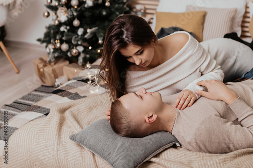 Lovely young romantic couple lying on bed garlands kissing hugging home cosy interior atmosphere New Year Christmas tree decorations holiday party celebrating concept winter evening 