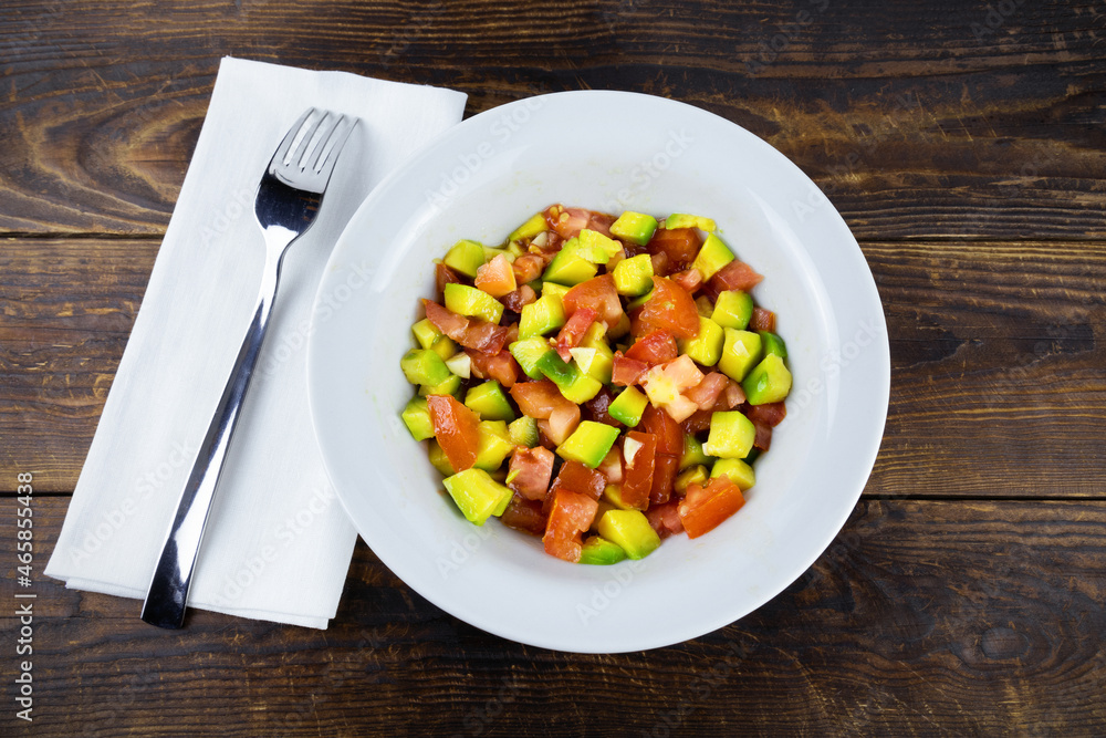 White plate with avocado and tomato salad, fork on napkin on brown wooden surface, top view, flatlay White plate with avocado and tomato salad, fork on napkin on brown wooden surface, top view, flatlay