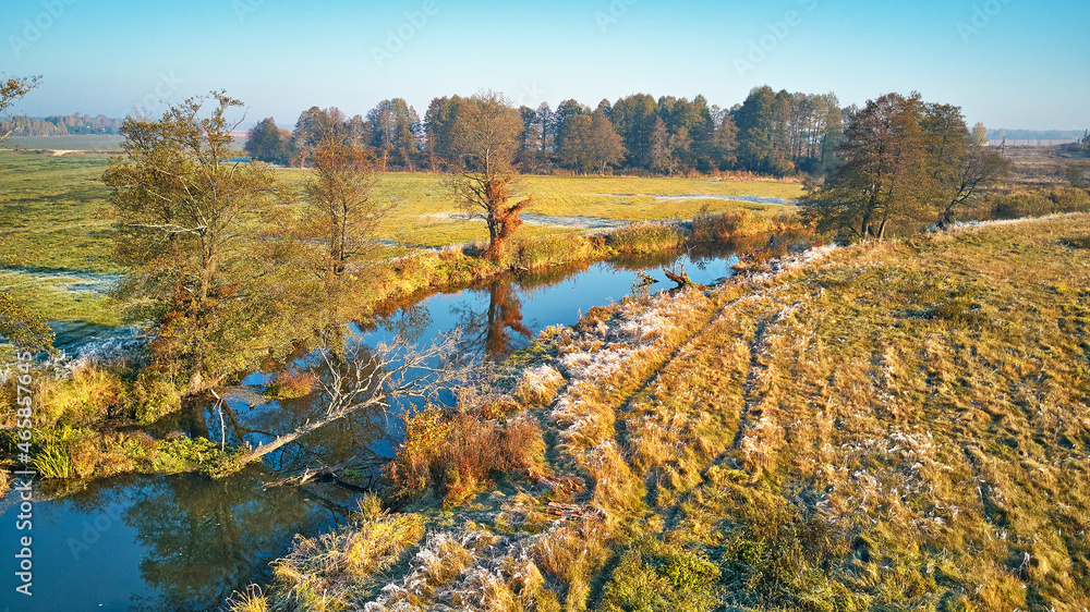 Autumn rural landscape. Frost on grass. River, green field, meadow, trees on riverbank. Dirt road on grassland.