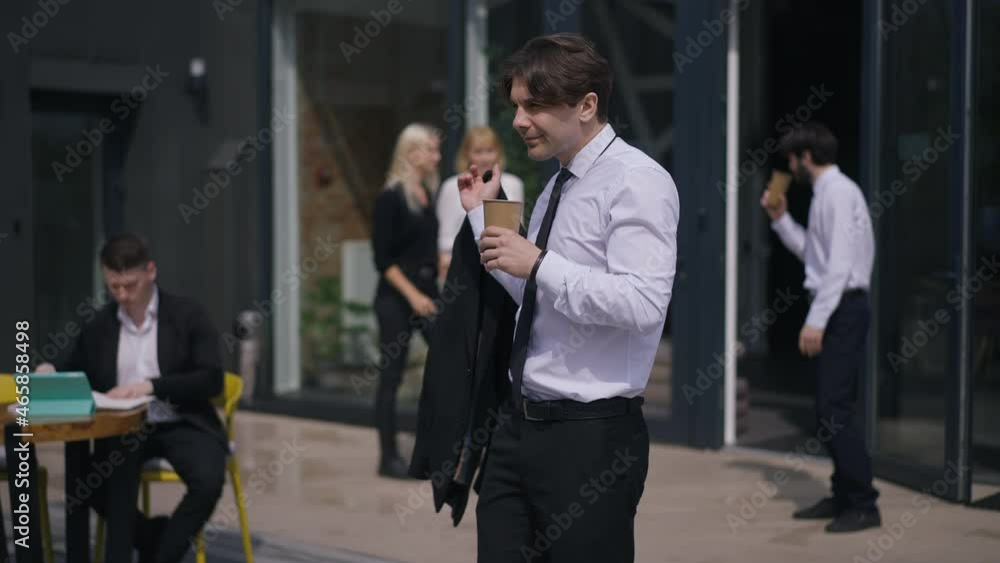 Confident positive businessman drinking coffee outdoors on terrace thinking with blurred colleagues talking discussing planning at background on sunny day. Confidence and business lifestyle Confident positive businessman drinking coffee outdoors on terrace thinking with blurred colleagues talking discussing planning at background on sunny day. Confidence and business lifestyle