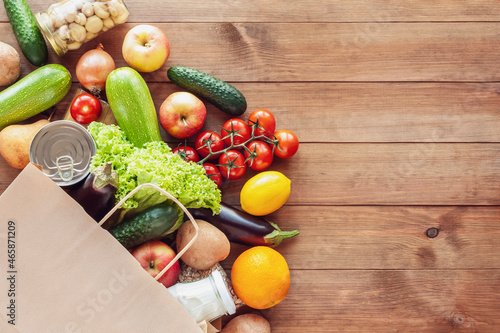Paper grocery bag with fresh vegetables, fruits, milk and canned goods on wooden backdrop. Food delivery, shopping, donation concept. Healthy food background. Flat lay, copy space.