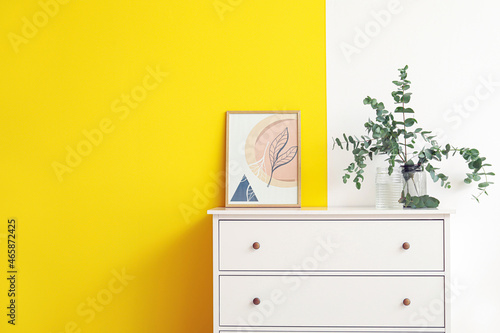 Vase with eucalyptus branches and picture on chest of drawers near color wall © Pixel-Shot
