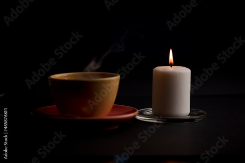candle lit with flame  with cup of tea with smoke  black background.  focus on candle .