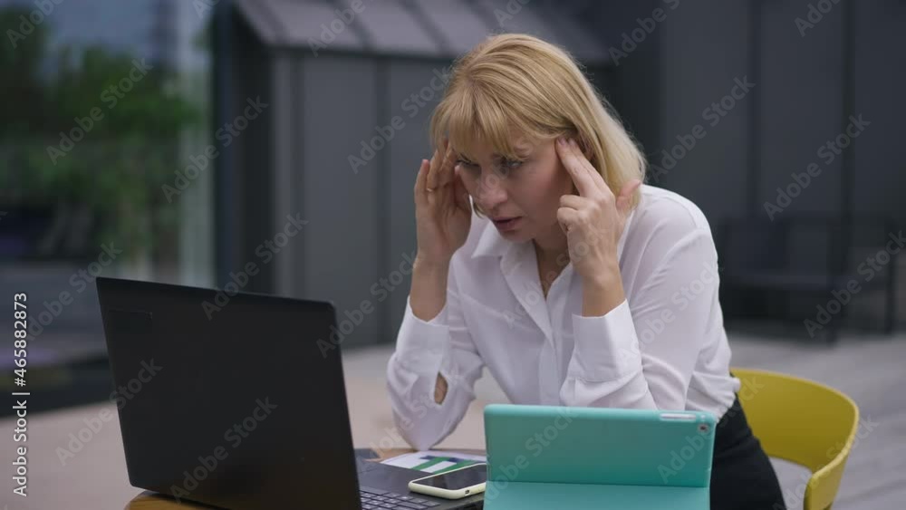 Overwhelmed beautiful woman rubbing temples sitting with laptop on office terrace. Portrait of tired exhausted Caucasian female manager planning strategy outdoors. Slow motion Overwhelmed beautiful woman rubbing temples sitting with laptop on office terrace. Portrait of tired exhausted Caucasian female manager planning strategy outdoors. Slow motion