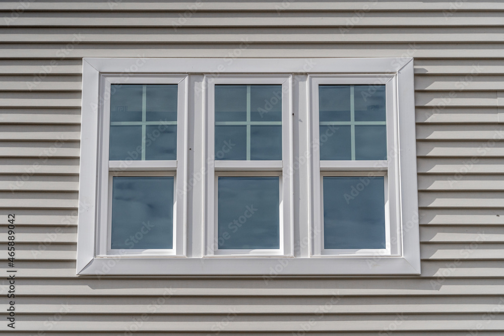 Triple hung window with fixed top sash and bottom sash that slides up, sash divided by white grilles a surrounded by white elegant frame  horizontal white vinyl siding on a new construction residence