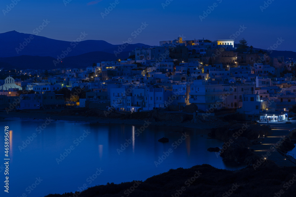 Blue night over a mountainous Greek island with a white city. Street lights are reflected in the calm sea