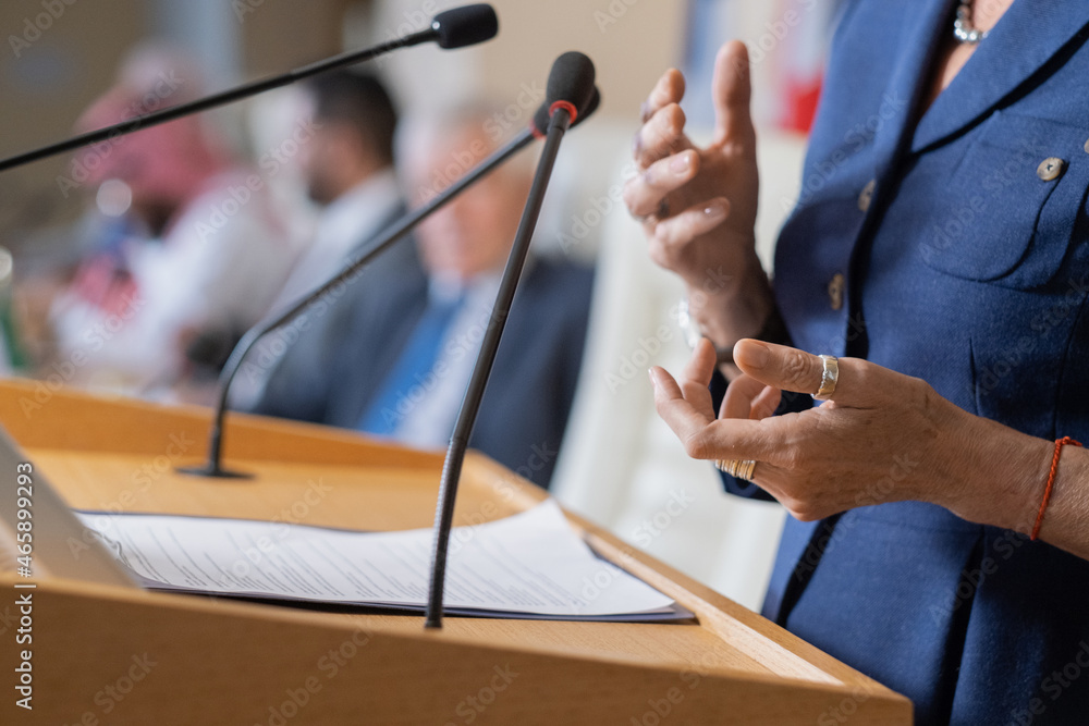 Close-up of unrecognizable female politician in rings standing at rostrum with microphones and clipboard while addressing conference - obrazy, fototapety, plakaty 