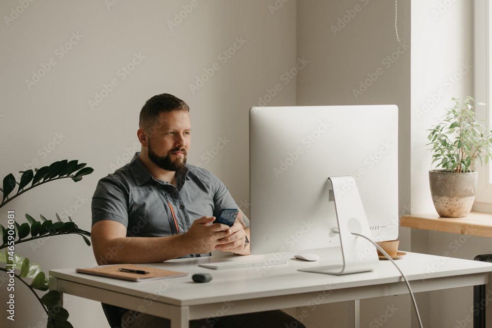 A smiling man works remotely on a desktop computer. A guy with a beard is holding a cellphone during a report of a colleague at a video conference at home. A teacher is preparing for an online lecture