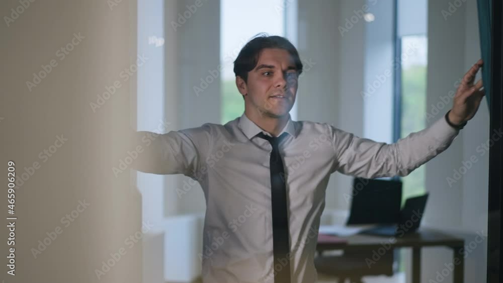 Young Caucasian man in white shirt and black necktie opening curtains in new office in slow motion looking out window thinking and walking to table. Positive happy employee indoors at workplace Young Caucasian man in white shirt and black necktie opening curtains in new office in slow motion looking out window thinking and walking to table. Positive happy employee indoors at workplace
