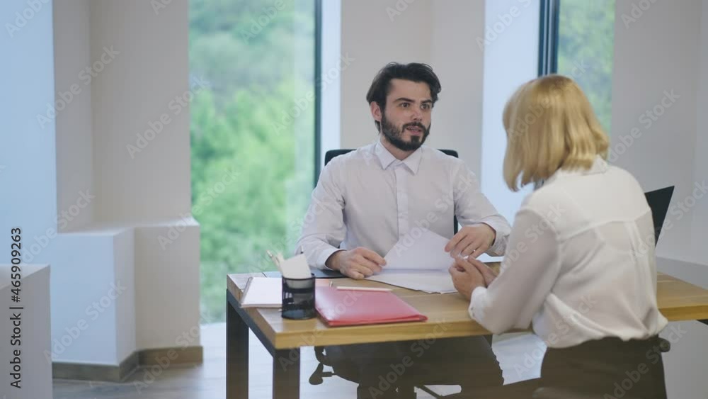 Bearded male manager sitting in office with female client passing document for sign talking in slow motion. Handsome positive Caucasian man serving customer indoors Bearded male manager sitting in office with female client passing document for sign talking in slow motion. Handsome positive Caucasian man serving customer indoors