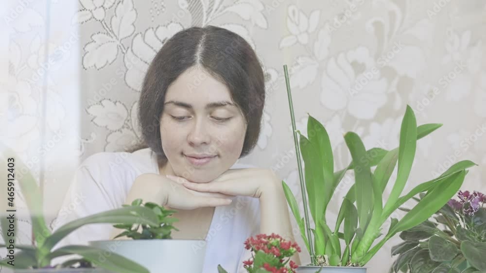 Portrait of a smiling woman inspecting a flower garden on the windowsill. The concept of home cultivation of flowers and plants. Portrait of a smiling woman inspecting a flower garden on the windowsill. The concept of home cultivation of flowers and plants.
