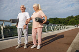 Joyful senior man and woman with rolled mats and earphones on footbridge