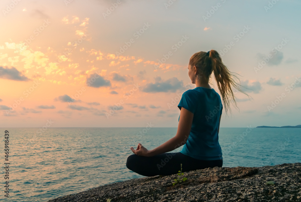 Young woman in lotus position meditates on rocks by the sea at sunrise. Colorful sky on the background.