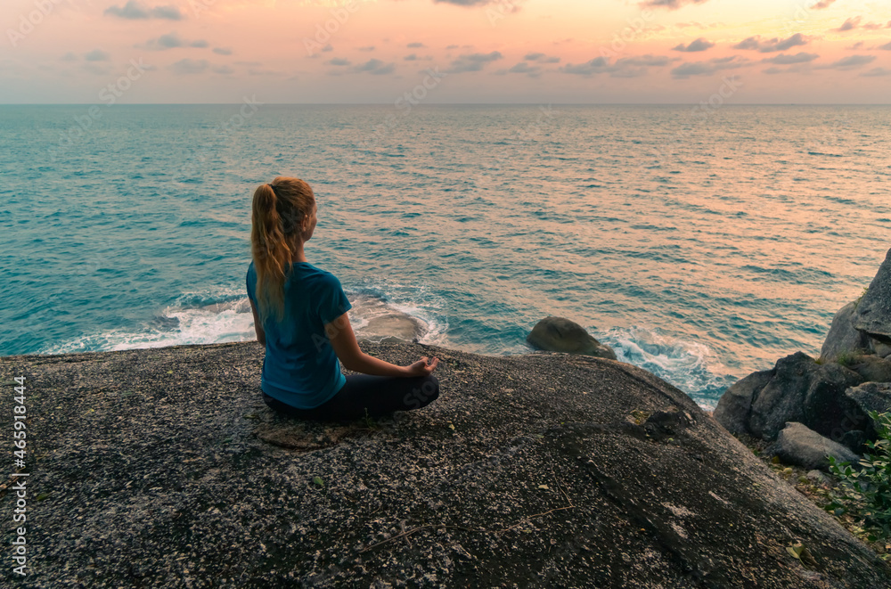 Young woman in lotus position meditates on rocks by the sea at sunrise. Colorful sky on the background. Young woman in lotus position meditates on rocks by the sea at sunrise. Colorful sky on the background.