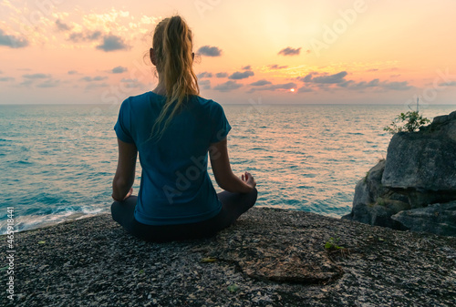 Young woman in lotus position meditates on rocks by the sea at sunrise. Colorful sky on the background.