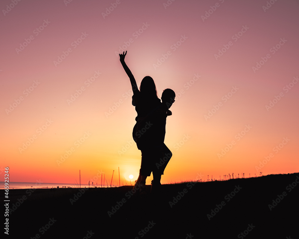 Couple en silhouette, amoureux. Un couché de soleil derrière le couple, elle est portée par son petit ami et fait coucou avec sa main.