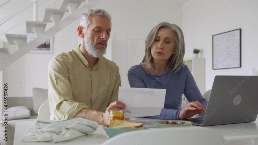 Senior middle aged couple holding financial documents paper bills paying bank loan online, calculating pension fees, payments, taxes, planning family retirement money finances using laptop at home. Senior middle aged couple holding financial documents paper bills paying bank loan online, calculating pension fees, payments, taxes, planning family retirement money finances using laptop at home.