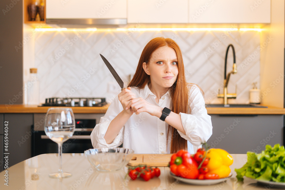 Portrait of attractive redhead young woman with mad eyes holding big knife sitting at table with cutting board and looking at camera in light kitchen room with modern interior, close-up.