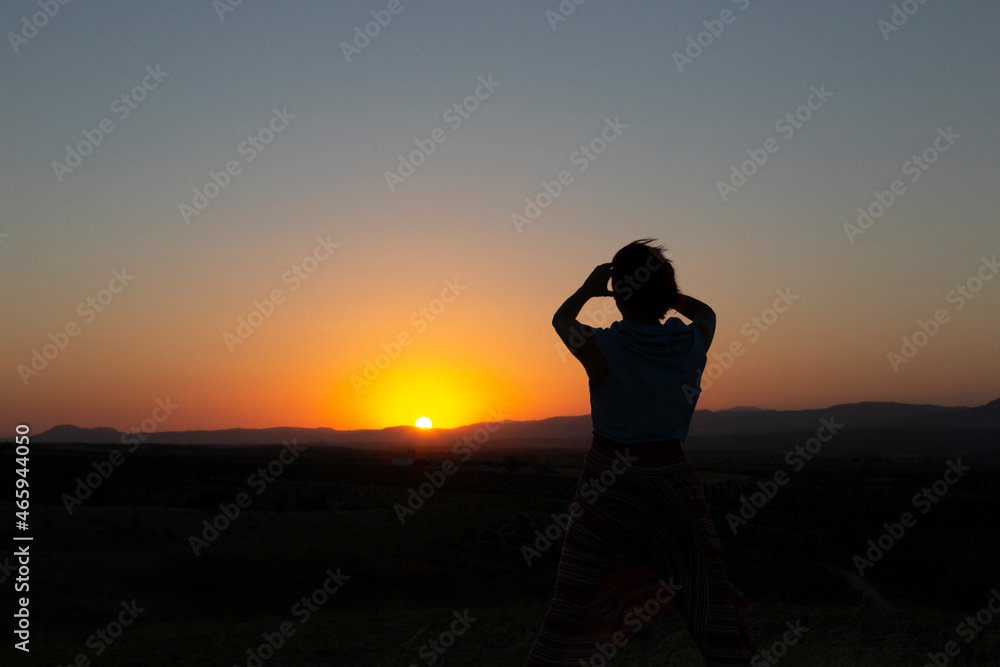 Fototapeta premium Silueta de mujer con los brazos levantados al caer la tarde, puesta de sol