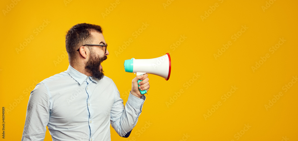 Horizontal photo of a handsome man wearing blue shirt and eyeglasses standing isolated over yellow background, screaming in loudspeaker, empty space Horizontal photo of a handsome man wearing blue shirt and eyeglasses standing isolated over yellow background, screaming in loudspeaker, empty space
