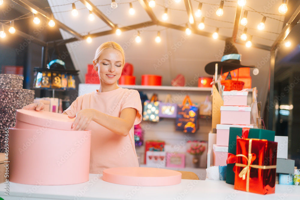 Obraz premium Attractive cheerful female seller sorting out holiday boxes standing behind store counter preparing to wrap Christmas presents at mall. Concept of new year, preparing xmas gifts.