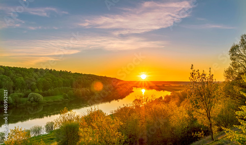 Scenic view at beautiful spring sunset on a shiny river valley with green branches, trees, bushes, grass, golden sun, calm water ,deep blue cloudy sky and forest on a background, spring landscape