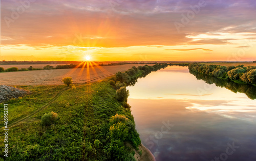 Scenic view at beautiful summer river sunset with reflection on water with green bushes  grass  golden sun rays  calm water  deep cloudy sky and glow on a background  spring   evening landscape