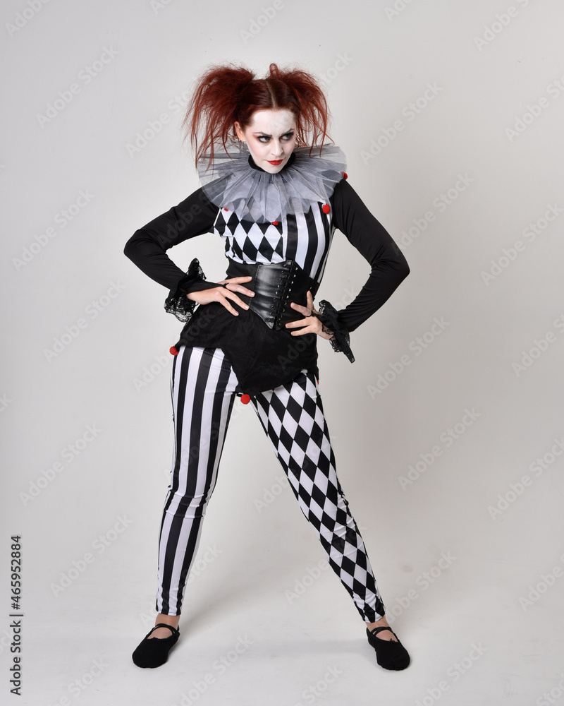 Full length portrait of red haired girl wearing a black and white clown jester costume, theatrical circus character. Standing pose isolated on studio background. Full length portrait of red haired girl wearing a black and white clown jester costume, theatrical circus character. Standing pose isolated on studio background.