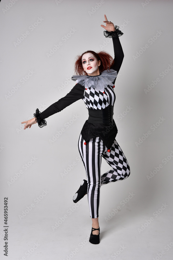 Obraz premium Full length portrait of red haired girl wearing a black and white clown jester costume, theatrical circus character. Standing pose isolated on studio background.