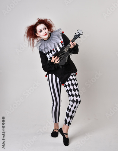 Full length portrait of red haired girl wearing a black and white clown jester costume, theatrical circus character. Standing pose isolated on studio background.