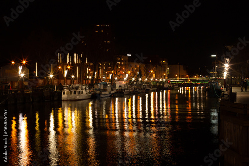 Klaipeda, Lithuania - 11 28 2020: boats at night. Beautiful city lights illumination reflecting in the water, boats and buildings, calm November street. A night city photo good for touristic booklets