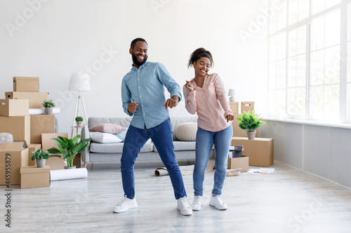 Smiling millennial african american husband and wife dancing together in room with cardboard boxes with stuff