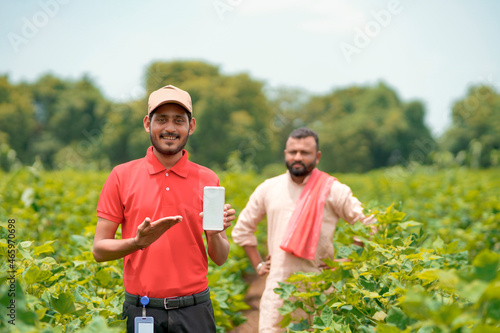 Young indian agronomist or banker showing smartphone with farmers at agriculture field.
