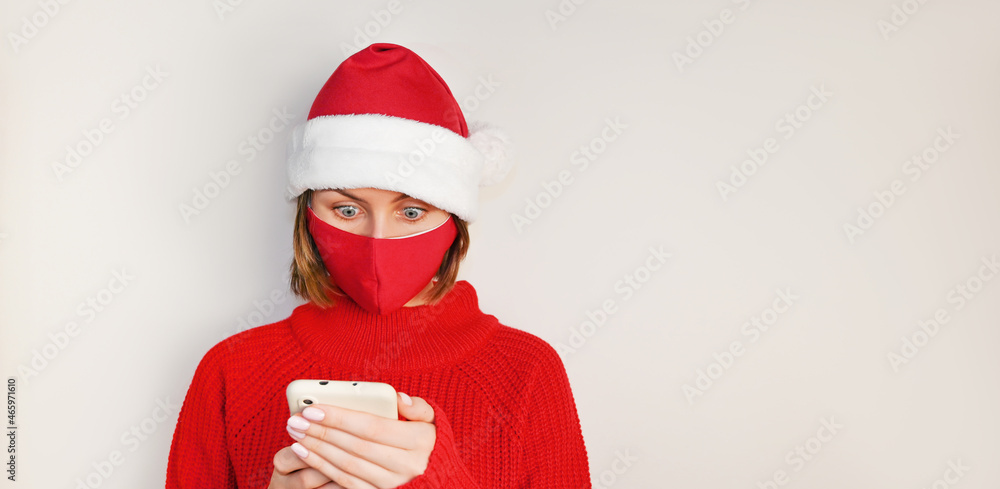 Christmas Woman in santa's hat, red sweater and wearing red medical nask stares surprised at phone's screen. Online shopping.Sale and discount concept.Mockup Christmas Woman in santa's hat, red sweater and wearing red medical nask stares surprised at phone's screen. Online shopping.Sale and discount concept.Mockup