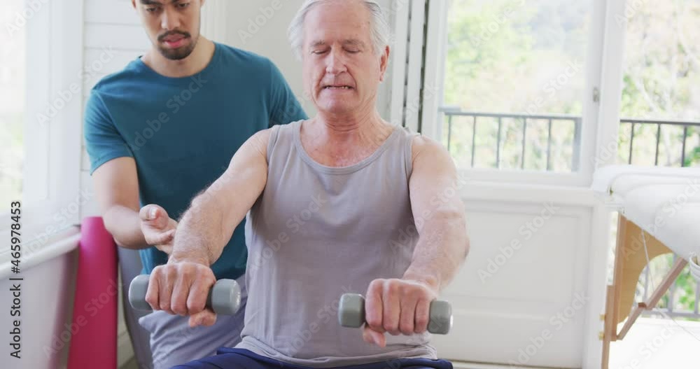 Retired senior man exercising with dumbbells by biracial male coach at fitness studio Retired senior man exercising with dumbbells by biracial male coach at fitness studio