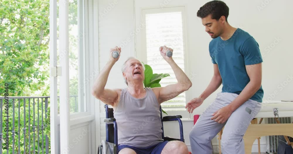Biracial male coach assisting disabled senior man exercising with dumbbells at fitness studio Biracial male coach assisting disabled senior man exercising with dumbbells at fitness studio