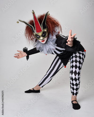 Full length portrait of red haired girl wearing a black and white clown jester costume, theatrical circus character. Sitting down on floor, isolated on studio background.