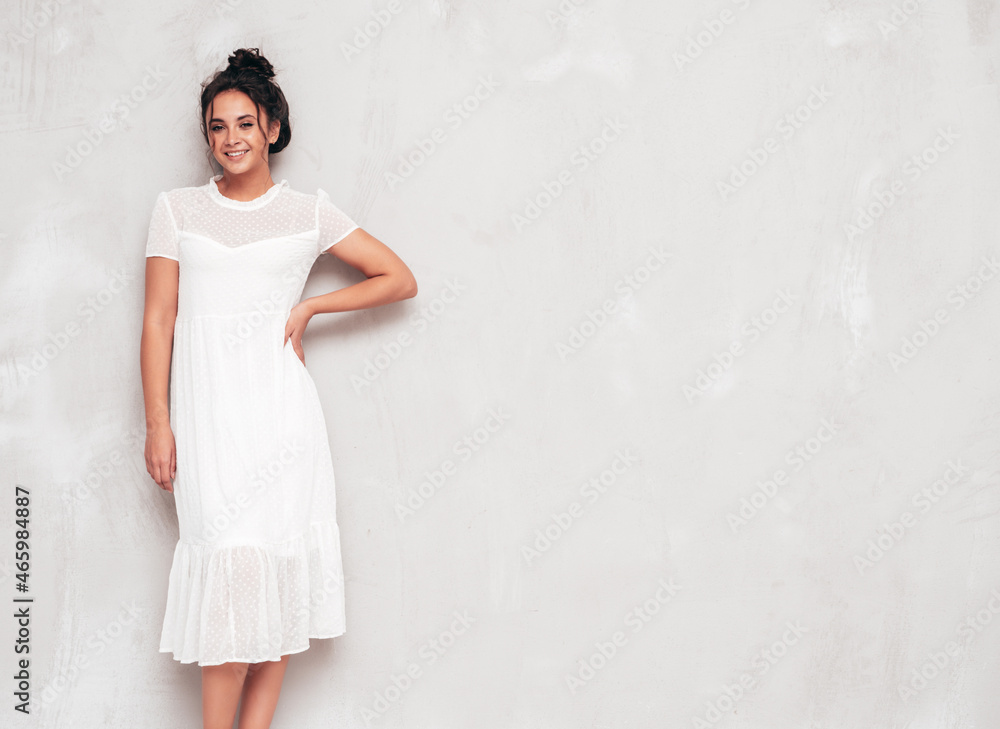 Portrait of young beautiful smiling female in trendy summer white dress. Sexy carefree woman posing near grey wall in studio. Positive model having fun indoors. Cheerful and happy Portrait of young beautiful smiling female in trendy summer white dress. Sexy carefree woman posing near grey wall in studio. Positive model having fun indoors. Cheerful and happy