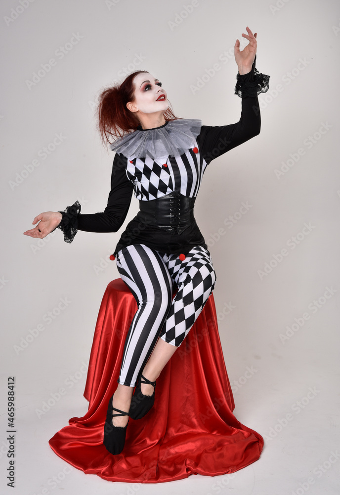 Full length portrait of red haired girl wearing a black and white clown jester costume, theatrical circus character. Sitting down on chair, isolated on studio background. Full length portrait of red haired girl wearing a black and white clown jester costume, theatrical circus character. Sitting down on chair, isolated on studio background.