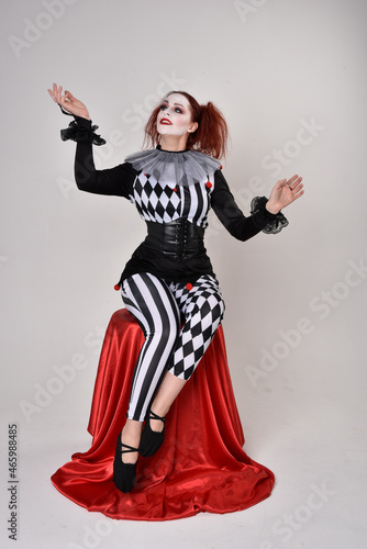 Full length portrait of red haired girl wearing a black and white clown jester costume, theatrical circus character. Sitting down on chair, isolated on studio background.